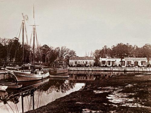 Historic black-and-white photo of a marina with sailboats and buildings along the water's edge, surrounded by trees.