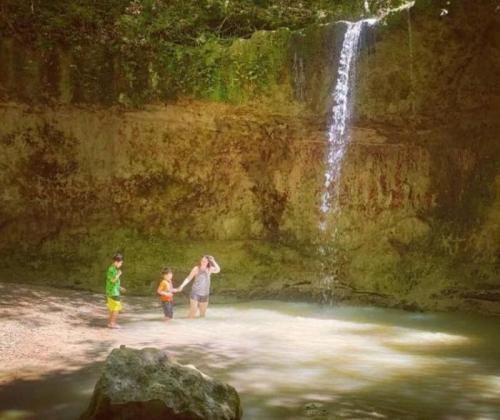A woman and two children play in a shallow pool beneath a waterfall surrounded by lush greenery.