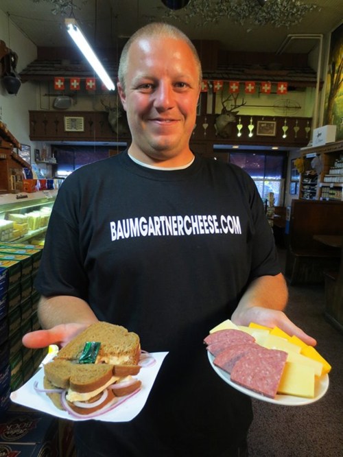 A man holds a sandwich and a plate of cheese and salami, wearing a shirt with "baumgartnercheese.com" printed on it.