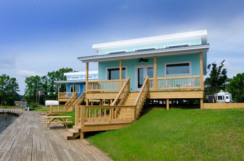 A colorful two-story house with a wooden deck, situated by a lake, surrounded by greenery and a wooden walkway.