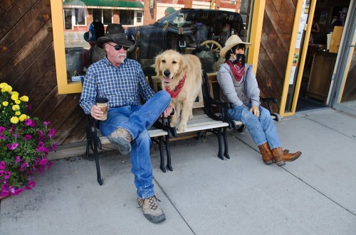 A man in a cowboy hat sits on a bench with a golden retriever, while a woman in a cowboy hat sits nearby.