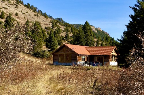 A rustic cabin with a red roof nestled among trees and mountains, surrounded by tall grass and shrubs.