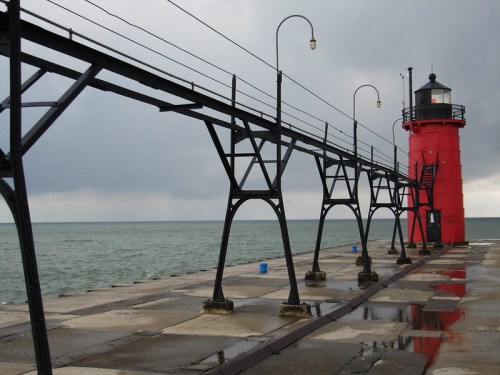 A red lighthouse stands at the end of a pier, with a cloudy sky and calm water in the background.