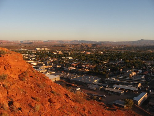 Aerial view of a town nestled in a valley, surrounded by red rock formations and distant mountains at sunset.