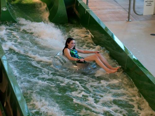 A girl rides a clear inner tube down a water slide, surrounded by splashing water in an indoor water park.