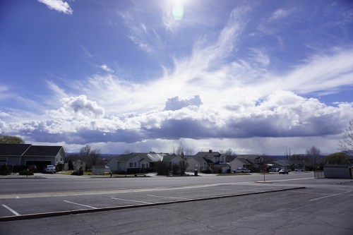 A wide view of a neighborhood under a bright blue sky with fluffy clouds and distant mountains.
