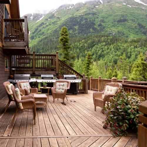 A wooden deck with cozy seating and grills, surrounded by lush green mountains and trees.