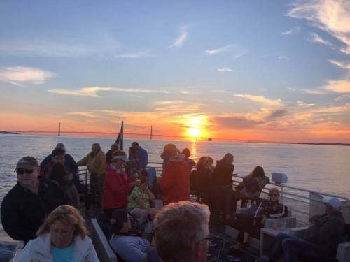 People enjoying a sunset cruise on a boat, with a bridge visible in the background and vibrant colors in the sky.