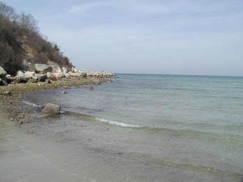 A serene beach scene with clear water, rocky shore, and gentle waves under a blue sky.