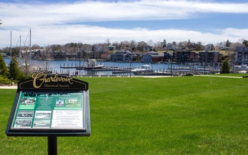 A green park with a sign in the foreground, overlooking a marina and waterfront homes in Charlevoix, Michigan.