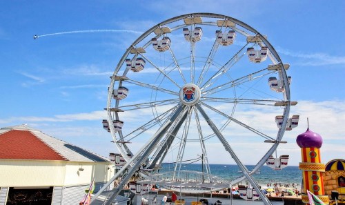 A large Ferris wheel stands by the beach, with an airplane flying overhead against a blue sky.
