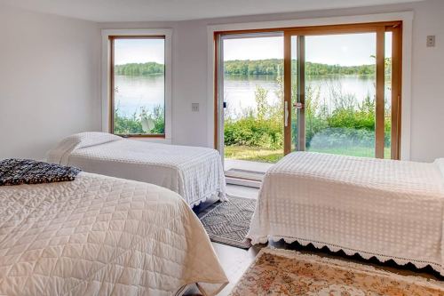 Cozy bedroom with two beds, large windows overlooking a serene lake and greenery outside.