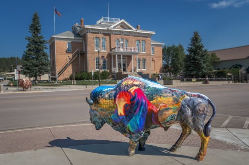 A colorful bison statue in front of a historic brick building with trees and an American flag in the background.
