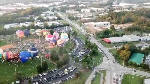 Aerial view of a hot air balloon festival with colorful balloons, a crowded parking area, and surrounding greenery.