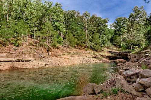A serene riverbank surrounded by lush green trees and rocky terrain under a clear blue sky.