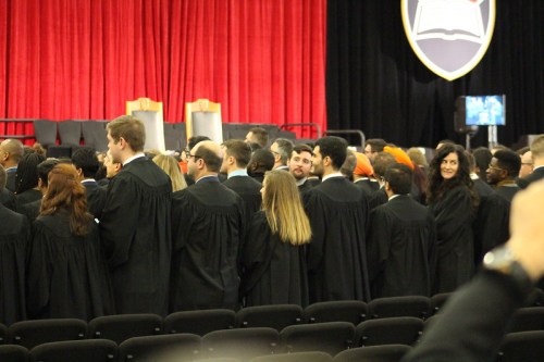 A group of graduates in black gowns and caps, gathered together, facing a stage with red curtains in the background.