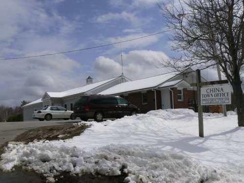 A snowy scene featuring a brick building with a sign reading "China Town Office" and parked cars nearby.