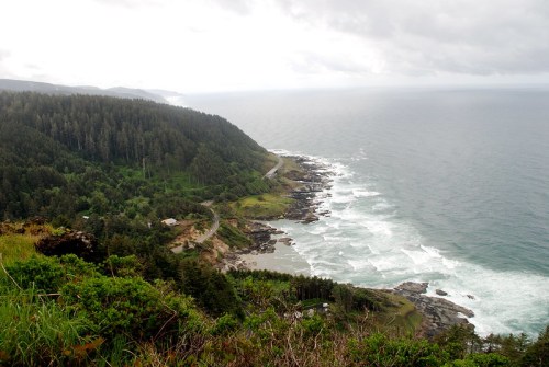 A coastal view featuring rugged cliffs, lush greenery, and waves crashing against the shore under a cloudy sky.