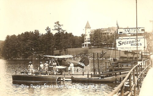 Historic photo of a lakeside dock with boats, a seaplane, and a hotel in the background, featuring people enjoying the scene.