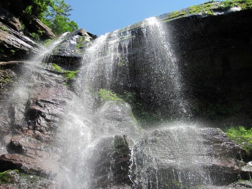 A cascading waterfall flows over rocky cliffs, surrounded by lush greenery under a clear blue sky.