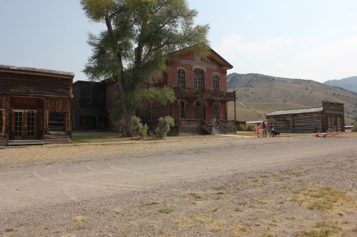 A dusty street in a ghost town with old wooden buildings and a large brick hotel, surrounded by mountains.