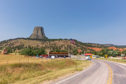 A scenic view of Devil's Tower in Wyoming, with a road, vehicles, and an American flag in the foreground.