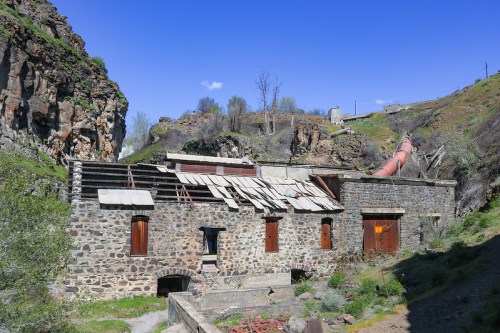 Abandoned stone building with a damaged roof, surrounded by green hills and rocky terrain under a clear blue sky.