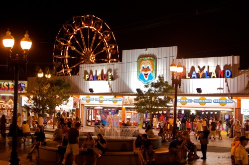 A vibrant amusement park at night featuring a clown sign, a fountain, and a large Ferris wheel in the background.
