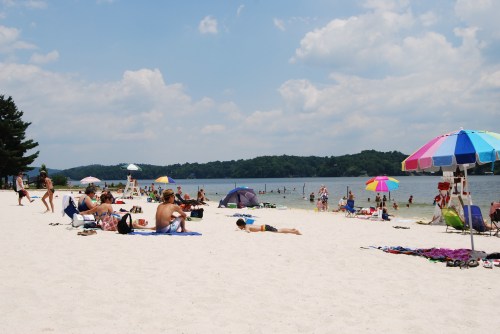 A sunny beach scene with people relaxing on the sand, colorful umbrellas, and a calm lake in the background.