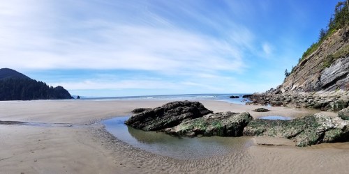 A panoramic view of a sandy beach with rocky formations, gentle waves, and a clear blue sky.