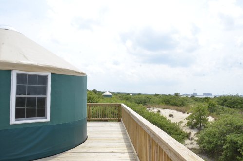 View of a green yurt on a wooden deck, surrounded by greenery and a cloudy sky in the background.