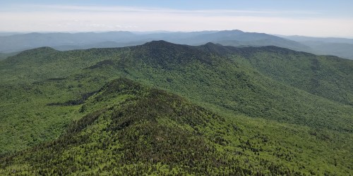 Aerial view of lush green mountains under a clear blue sky, showcasing rolling hills and distant peaks.