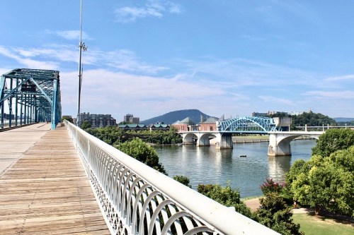 A scenic view of a blue bridge over a river, with buildings and mountains in the background under a clear sky.