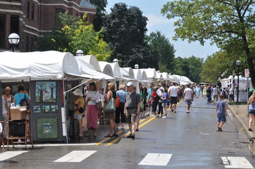 A bustling outdoor market with white tents, people browsing art, and a sunny day with trees in the background.