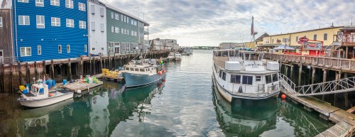 A scenic harbor with boats docked alongside colorful buildings and a cloudy sky reflecting on the water.