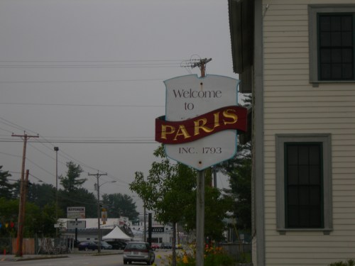 Sign reading "Welcome to Paris, INC. 1793" near a road, with a cloudy sky and buildings in the background.