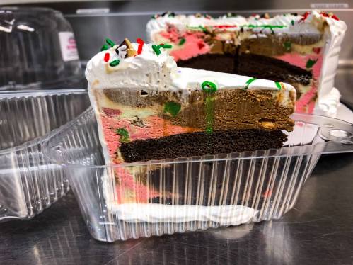 A slice of colorful layered cake with chocolate, pink, and green frosting, in a clear plastic container.