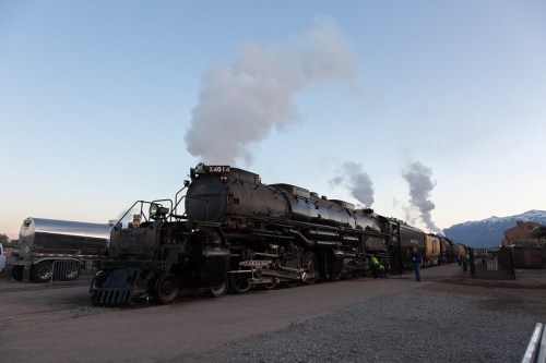 A vintage steam locomotive emits smoke as it stands on a track, with mountains in the background and workers nearby.