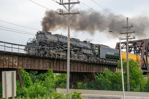 A steam locomotive crosses a bridge, emitting black smoke against a cloudy sky, surrounded by greenery.