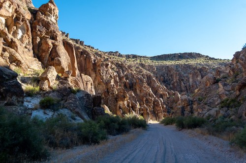 A dirt road winds through a rocky canyon with steep cliffs and sparse vegetation under a clear blue sky.