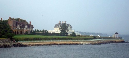 Coastal view featuring two large mansions and a gazebo along a rocky shoreline under a cloudy sky.
