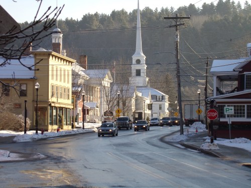 A snowy street scene in a small town, featuring a church steeple and cars driving along the road.