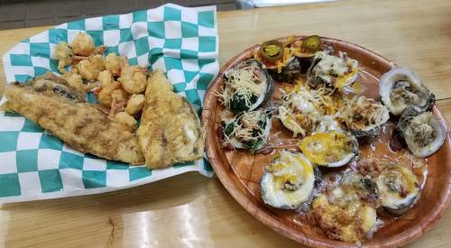 A wooden plate with baked oysters topped with cheese and jalapeños, alongside a basket of fried fish and shrimp.