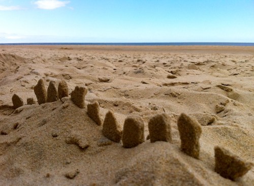 A sandy beach with small sand sculptures resembling a row of teeth against a clear blue sky and ocean in the background.