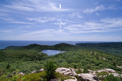A panoramic view of lush green hills, a serene lake, and the ocean under a bright blue sky with scattered clouds.