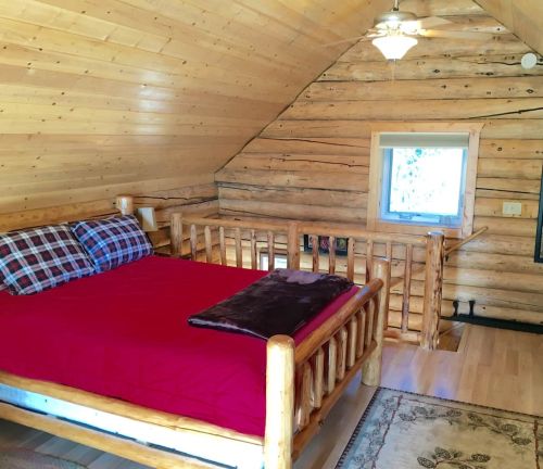 Cozy attic bedroom with a wooden log bed, plaid pillows, and a warm wooden interior. Natural light from a window.
