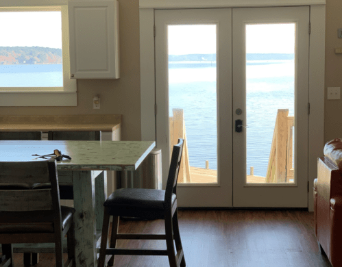 A cozy interior with a table and chairs, leading to a view of a lake through glass doors.