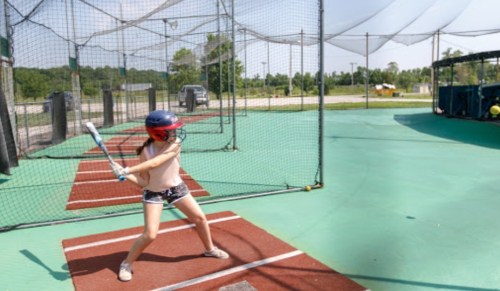 A young girl in a helmet prepares to swing a bat in a baseball batting cage on a sunny day.