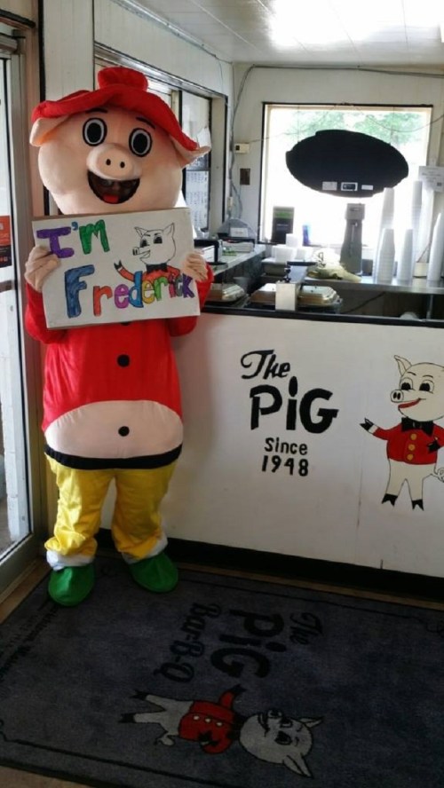 A person in a pig costume holds a sign saying "I'm Frederick" next to a counter with "The Pig Since 1948" logo.