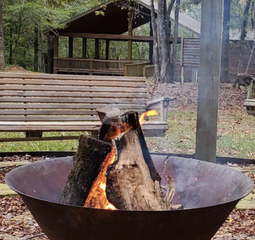 A fire burns in a metal fire pit, with smoke rising and wooden benches visible in a wooded area.